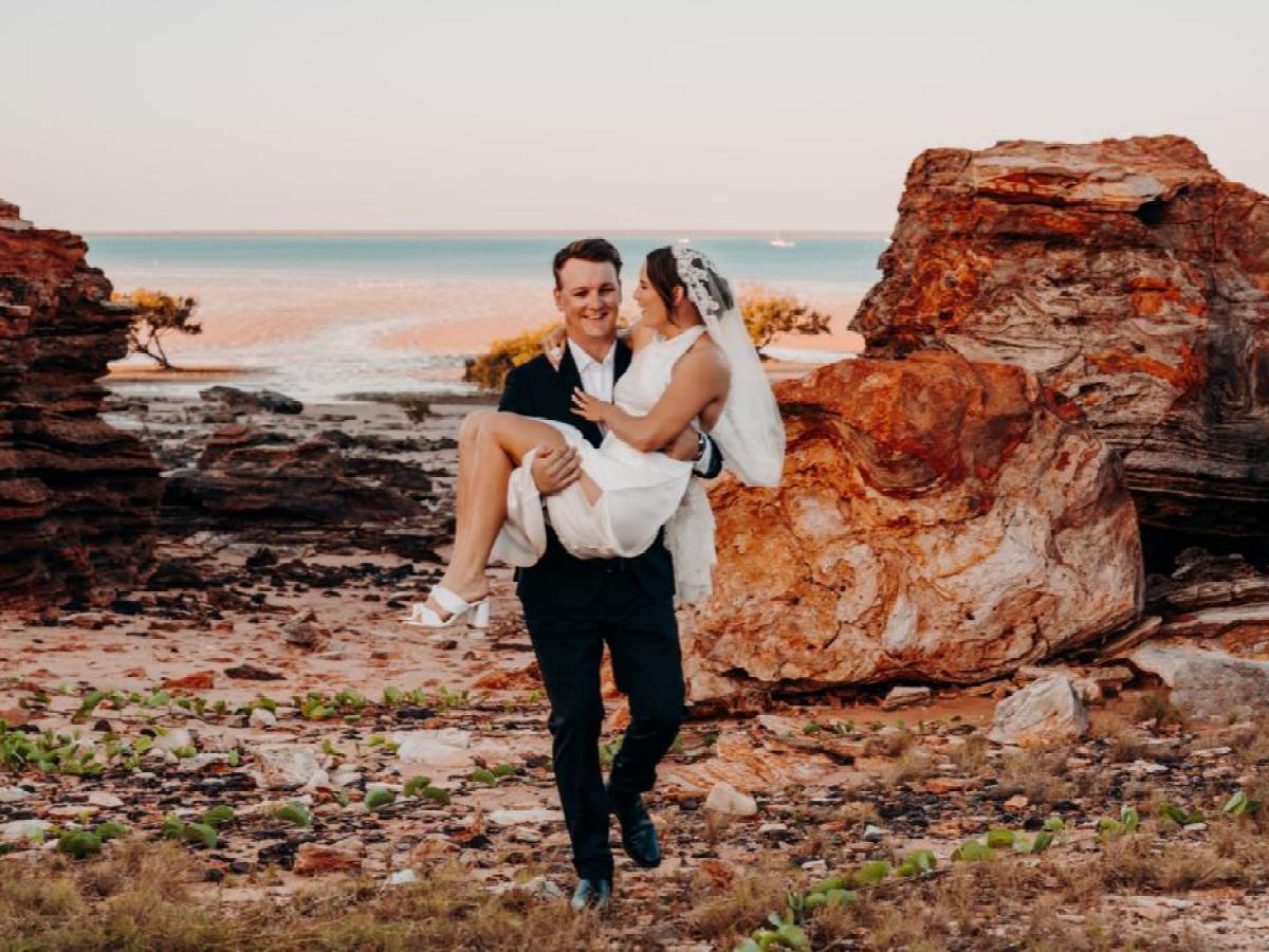 Groom carrying bride in rocky coastal landscape with ocean in background.
