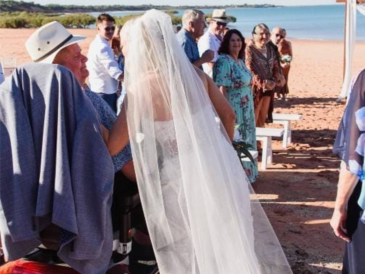 Bride in white dress walking on a beach aisle, guests seated and standing, clear sky and sea in the background.
